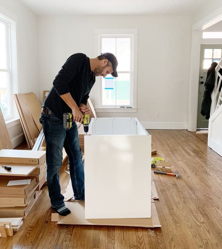 Installation in progress — fitter assembling a cabinet on site, hardwood floor, packing material and other cabinets stacked nearby